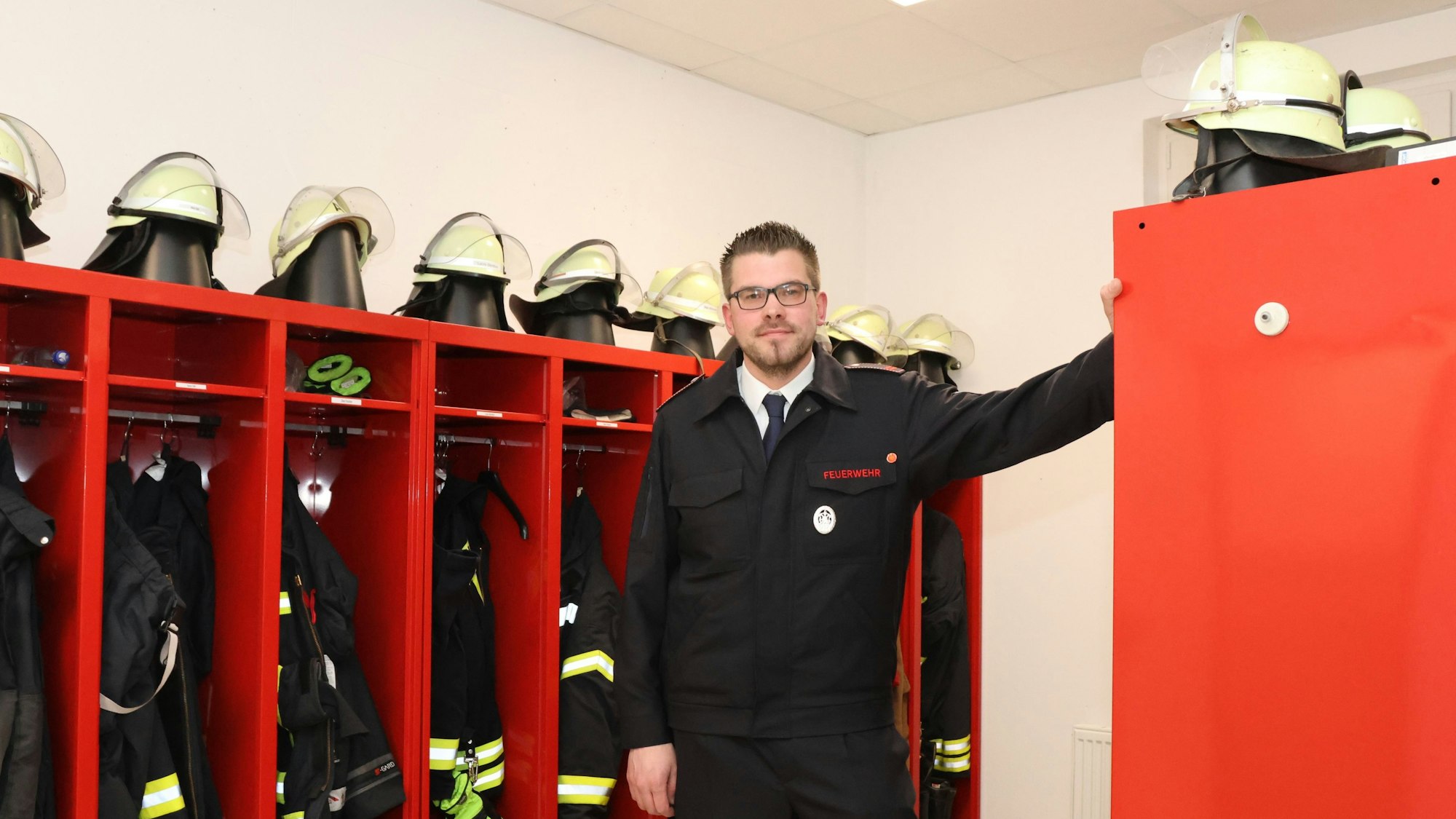 Ein Feuerwehrmann steht vor roten Spinden, in denen die Arbeitskleidung seiner Kameraden hängt. Auf den Spinden liegen Helme.
