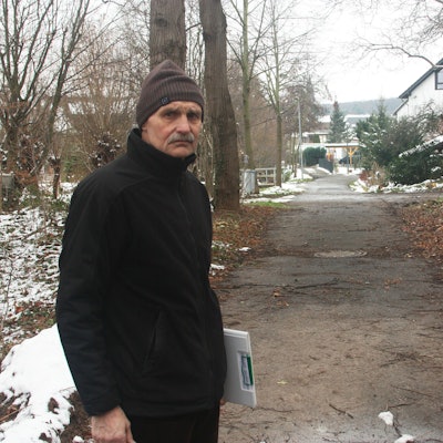 Blindert im Wald bei Schnee, hinter sich ein Weg, der auf den Ort zuführt.