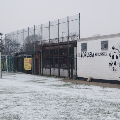 Der Rasen ist mit Schnee bedeckt, dahinter steht ein einstöckiges Vereinsheim mit der Aufschrift „FC Borussia Buir“.
