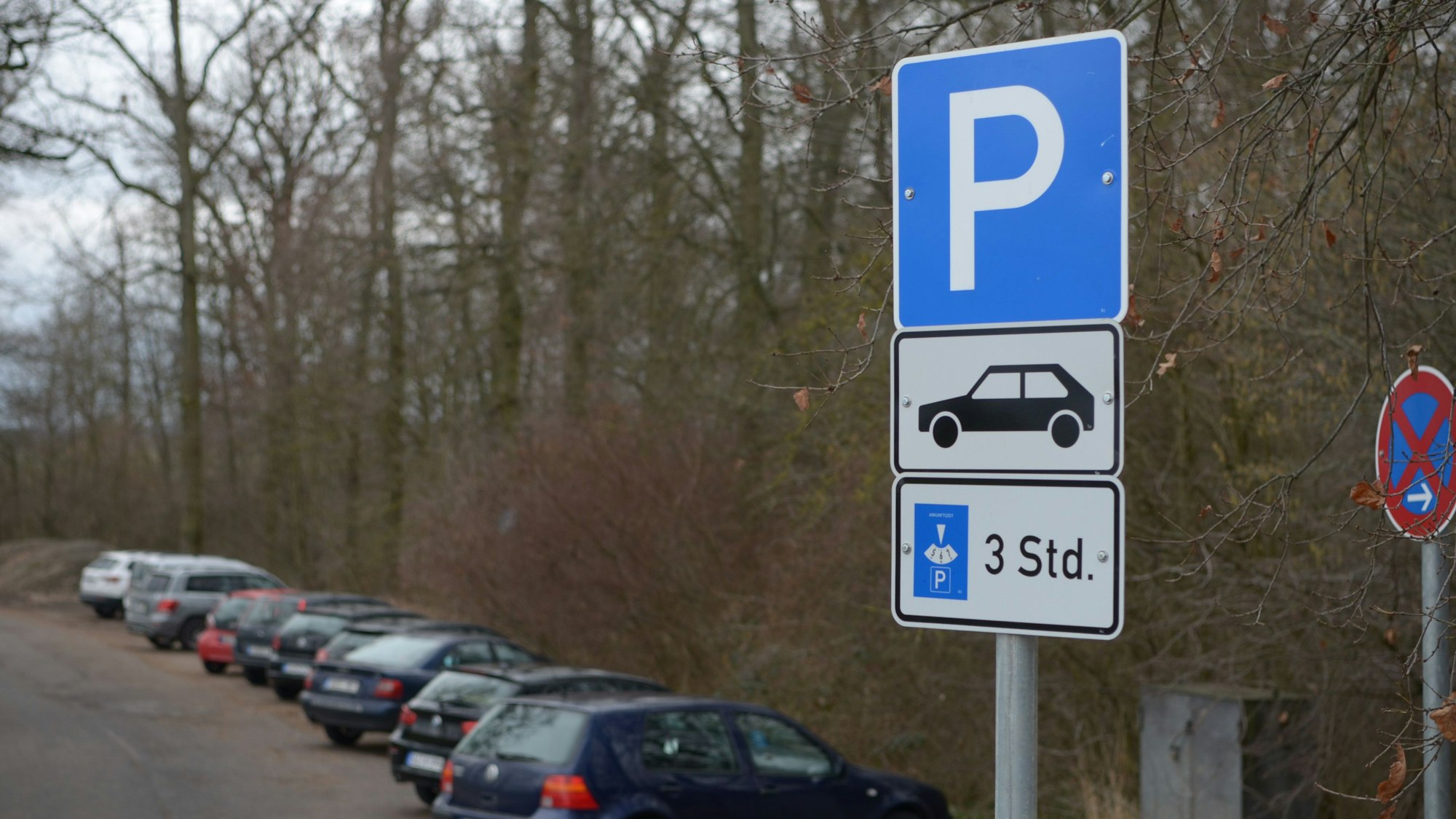 Man sieht im Vordergrund ein Verkehrsschild, im Hintergrund einen Parkplatz am Waldrand, auf dem Autos stehen.