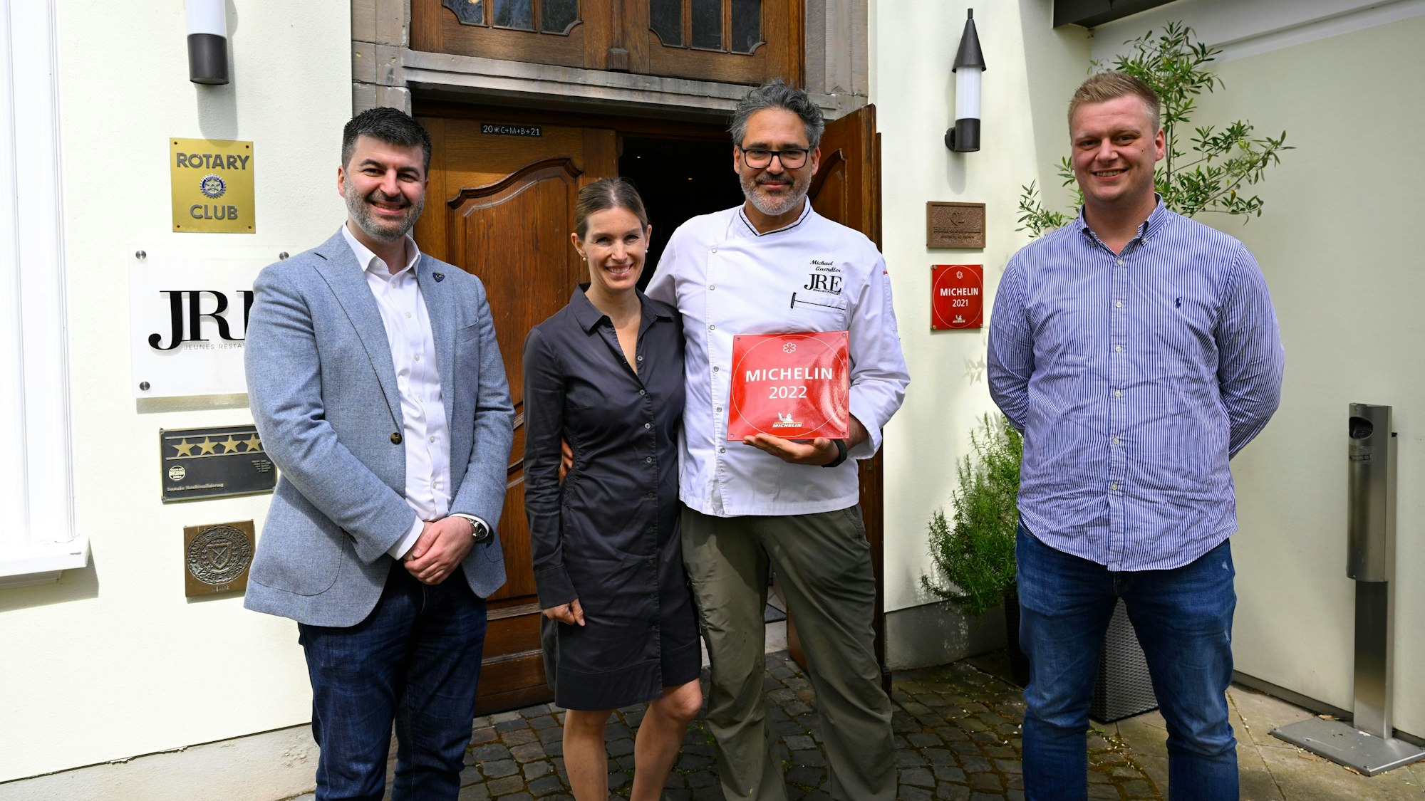Vier Personen stehen vor einem Gebäude mit einer großen Holztüre. Ein Mann hält eine rote Plakette mit der Michelin-Auszeichnung in der Hand.