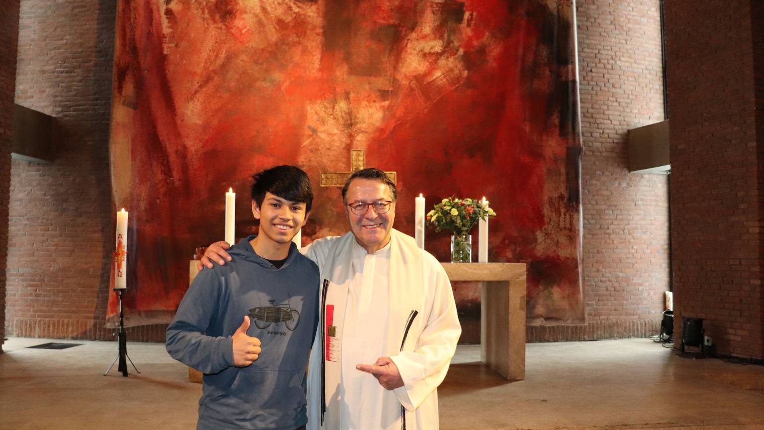 Die beiden stehen vor dem Altar. Hinter dem Altar hängt ein großes rötliches Tuch.