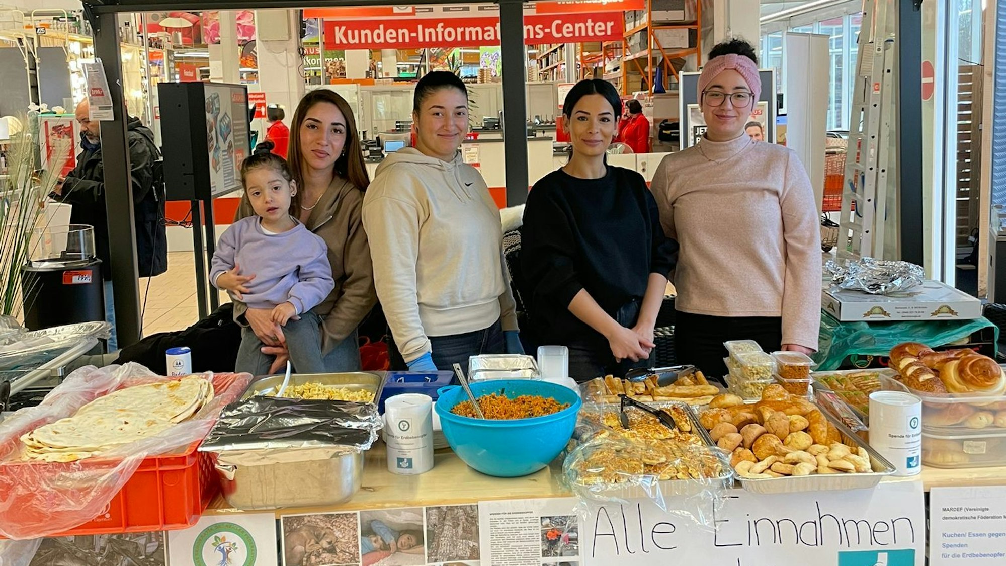 Hinter einem Marktstand stehen vier junge Frauen. Vor ihnen liegt Gebäck in der Auslage. Eine Frau hält ein kleines Mädchen auf dem Arm.