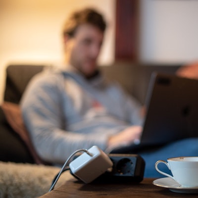 Ein Mann sitzt mit Laptop auf dem Sofa und macht seine Steuererklärung, vor ihm steht eine Tasse Tee.