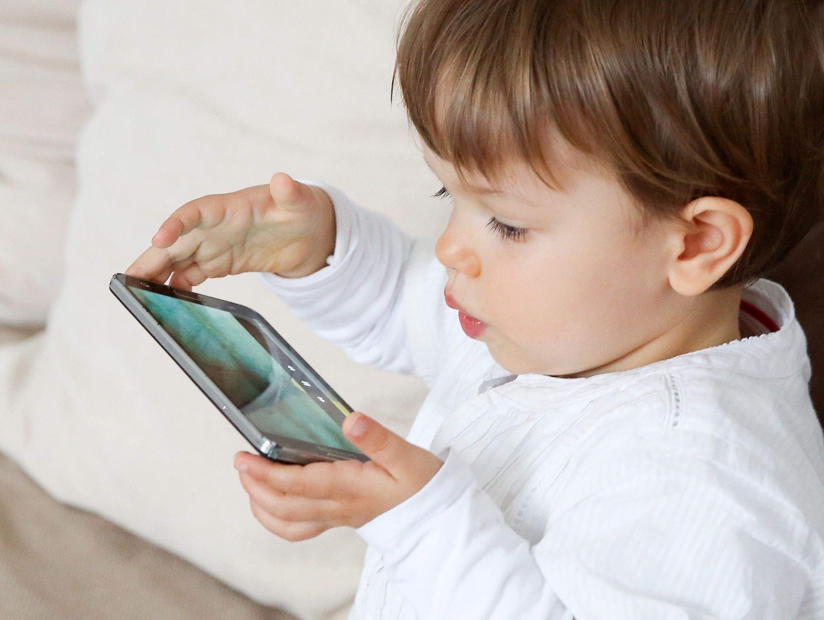 Ein Kleinkind sitzt auf der Couch und spielt mit einem Smartphone.