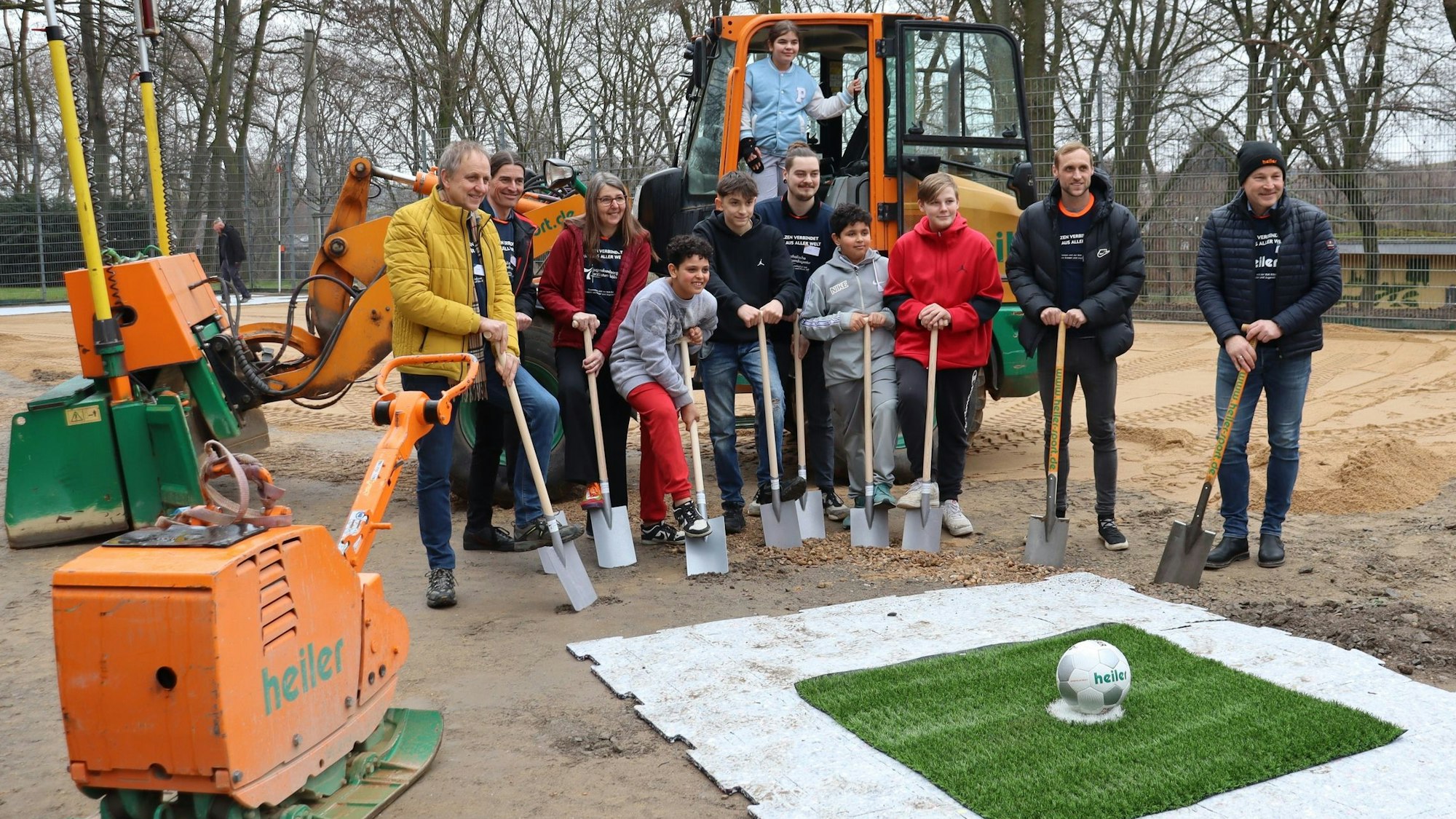 Marcel Risse mit Kindern und Schaufel in der Hand vor dem Kunstrasen, dahinter steht ein Bagger