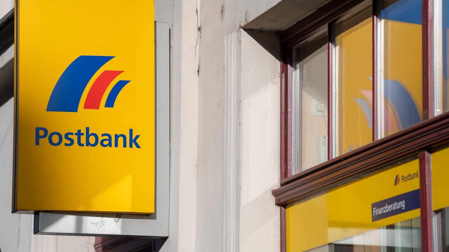 Ein Schild mit dem Logo der Postbank ist an der Fassade einer Bankfiliale angebracht.