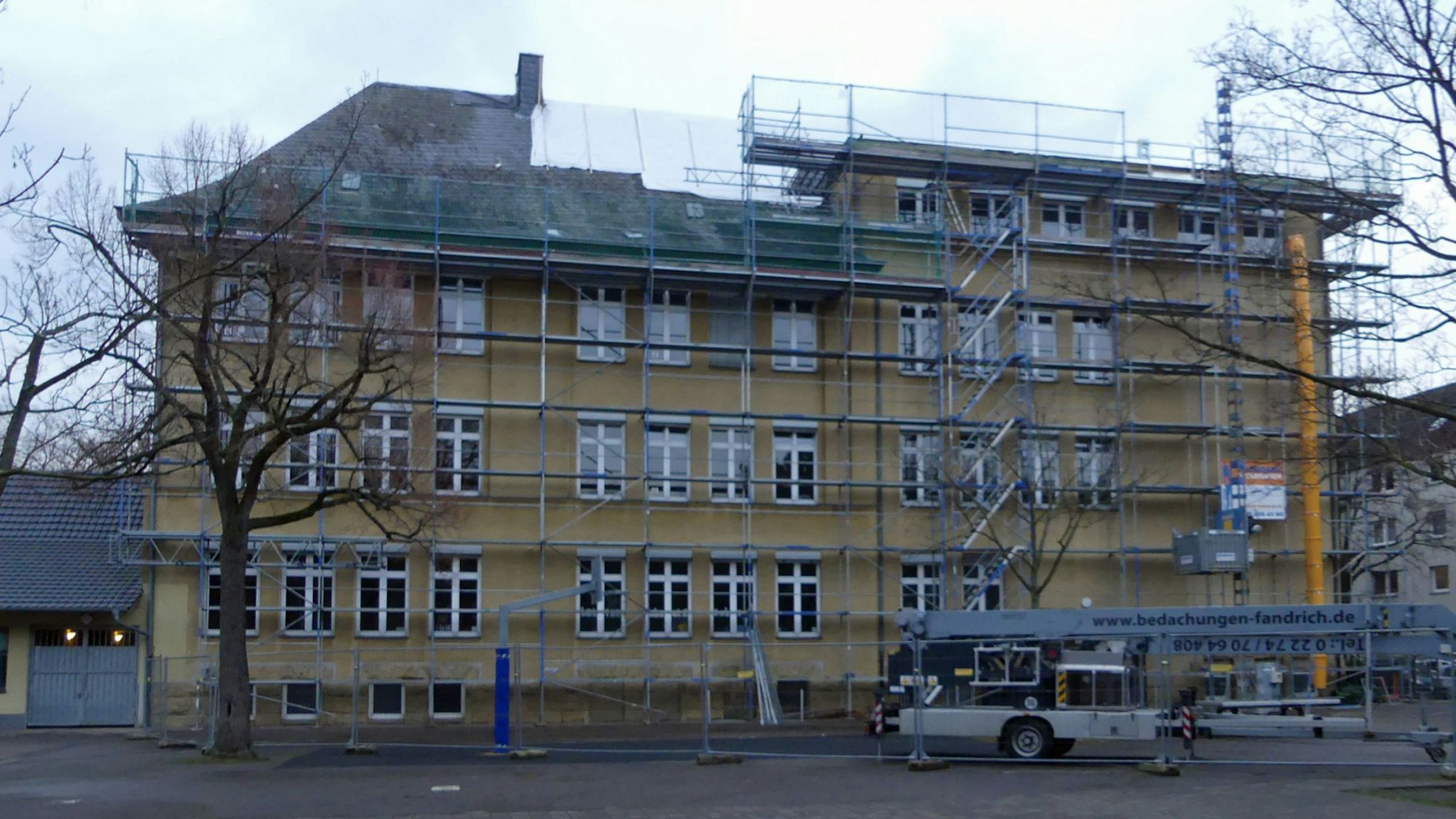 Ein mehrstöckiges großes Gebäude mit gelber Fassade ist zu sehen. Vor dem Haus stehen Gerüste, Teile des Dachs sind mit Planen zugedeckt.