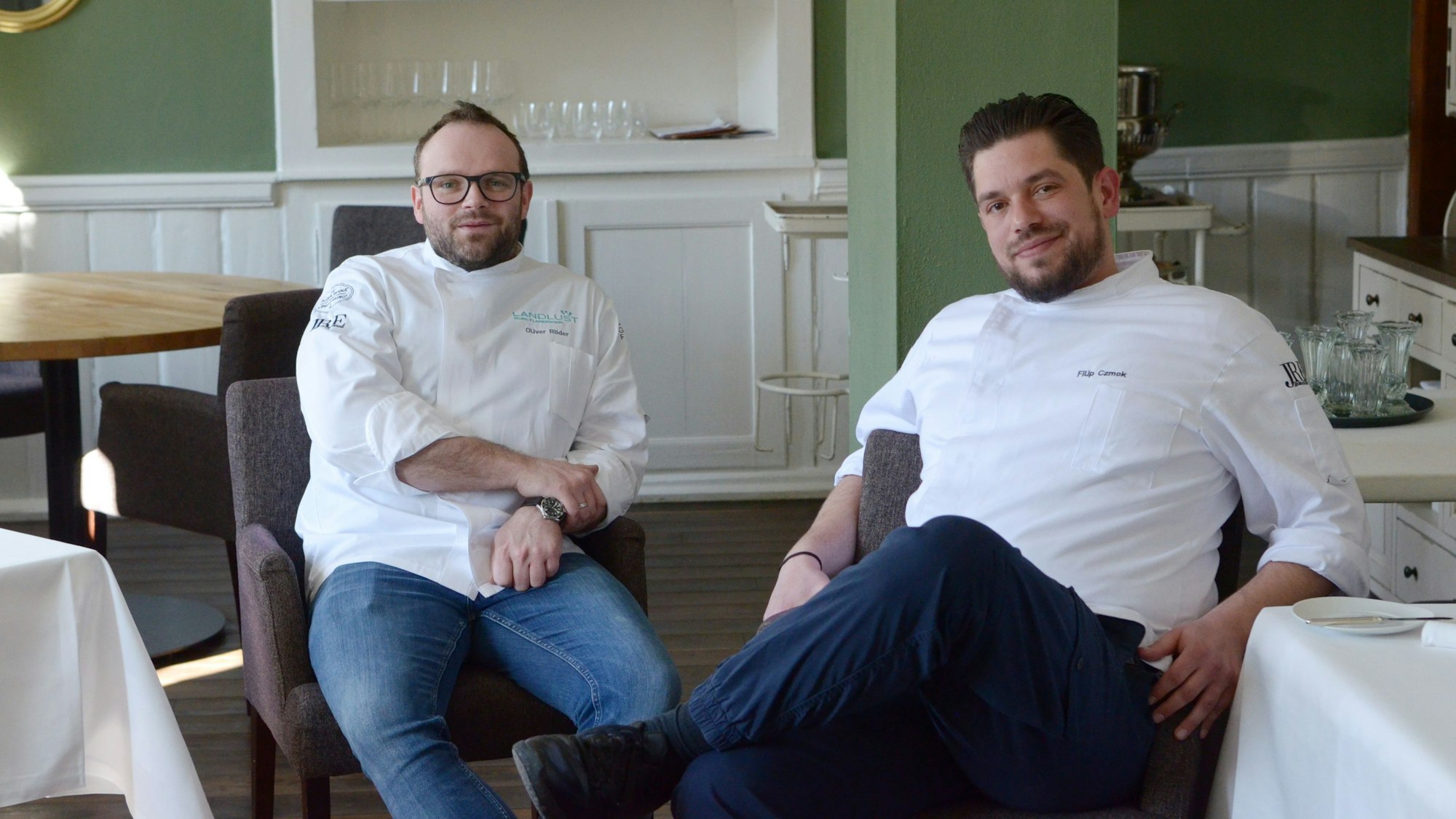 Der Geschäftsführer vom Bembergs Häuschen Oliver Röder und Küchenchef Filip Czmok sitzen in weißer Kochmontur im Gastraum ihres Restaurants.