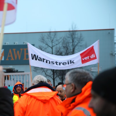 Streikende stehen mit einem Banner mit der Aufschrift „Warnstreik“ vor dem Werkstor der AWB Abfallwirtschaftsbetriebe.