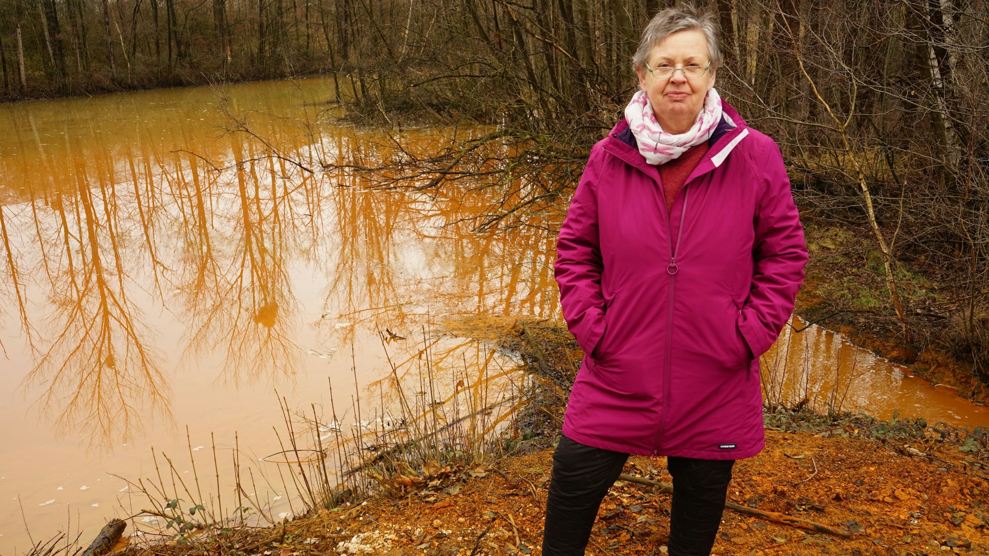 Auf dem Foto ist Naturschützerin Jutta Schnütgen-Weber zu sehen. Sie sorgt sich, dass belastetes Wasser aus dem Rückhaltebecken in die Erft fließt. Im Hintergrund ist das rot gefärbte Wasser zu sehen.