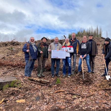 Mehrere Personen stehen mit Spaten auf einer kahlen Waldfläche nebeneinander. Vor ihnen sind Setzlinge zu sehen. In der Mitte halten sie einen Spendenscheck, auf dem die Summe 30.000 Euro eingetragen sind.