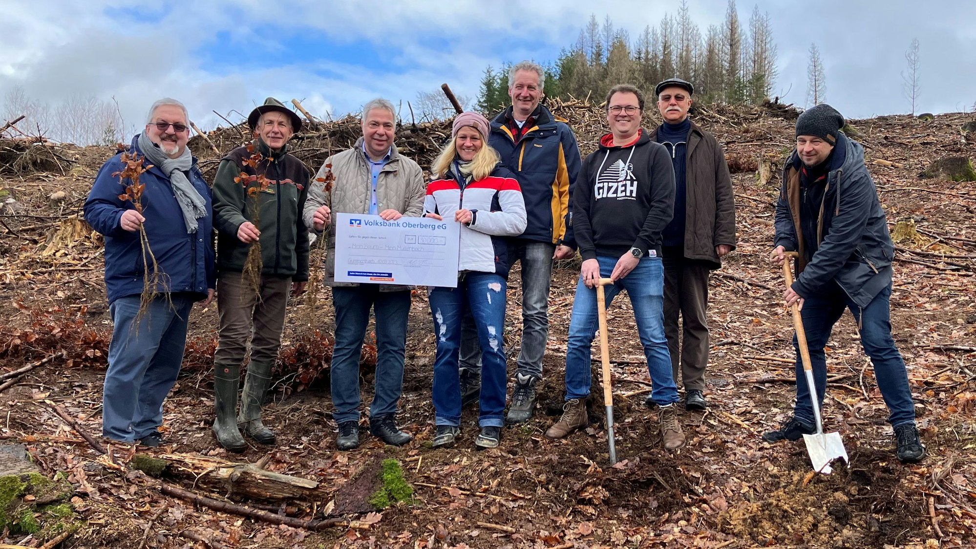Mehrere Personen stehen mit Spaten auf einer kahlen Waldfläche nebeneinander. Vor ihnen sind Setzlinge zu sehen. In der Mitte halten sie einen Spendenscheck, auf dem die Summe 30.000 Euro eingetragen sind.