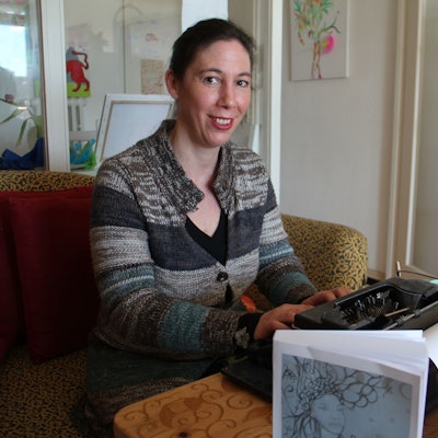 Nießen sitzt an einer Schreibmaschine mit ihrem Buch daneben und blickt in die Kamera.