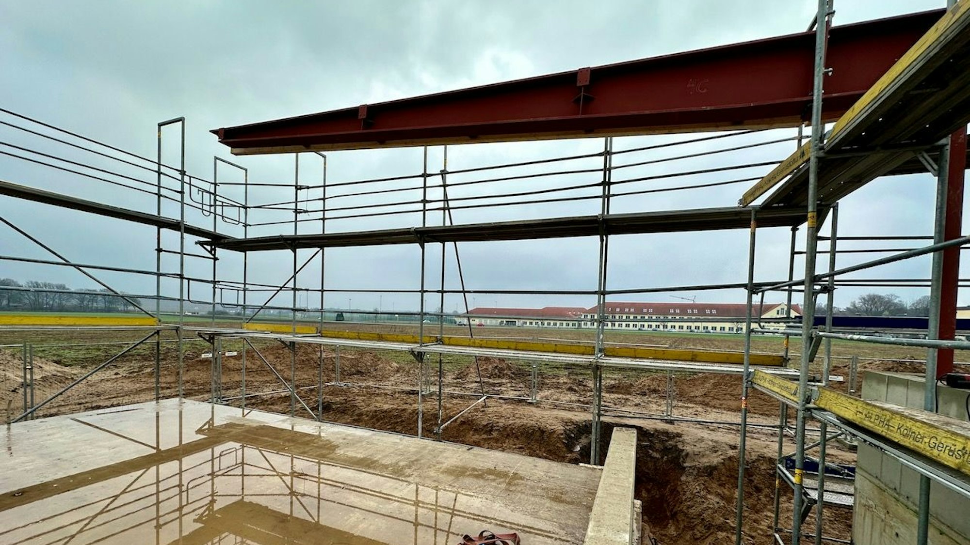 Ackerfläche mit aufgestelltem Gerüst. Flaches Altbau-Schulgebäude im Hintergrund