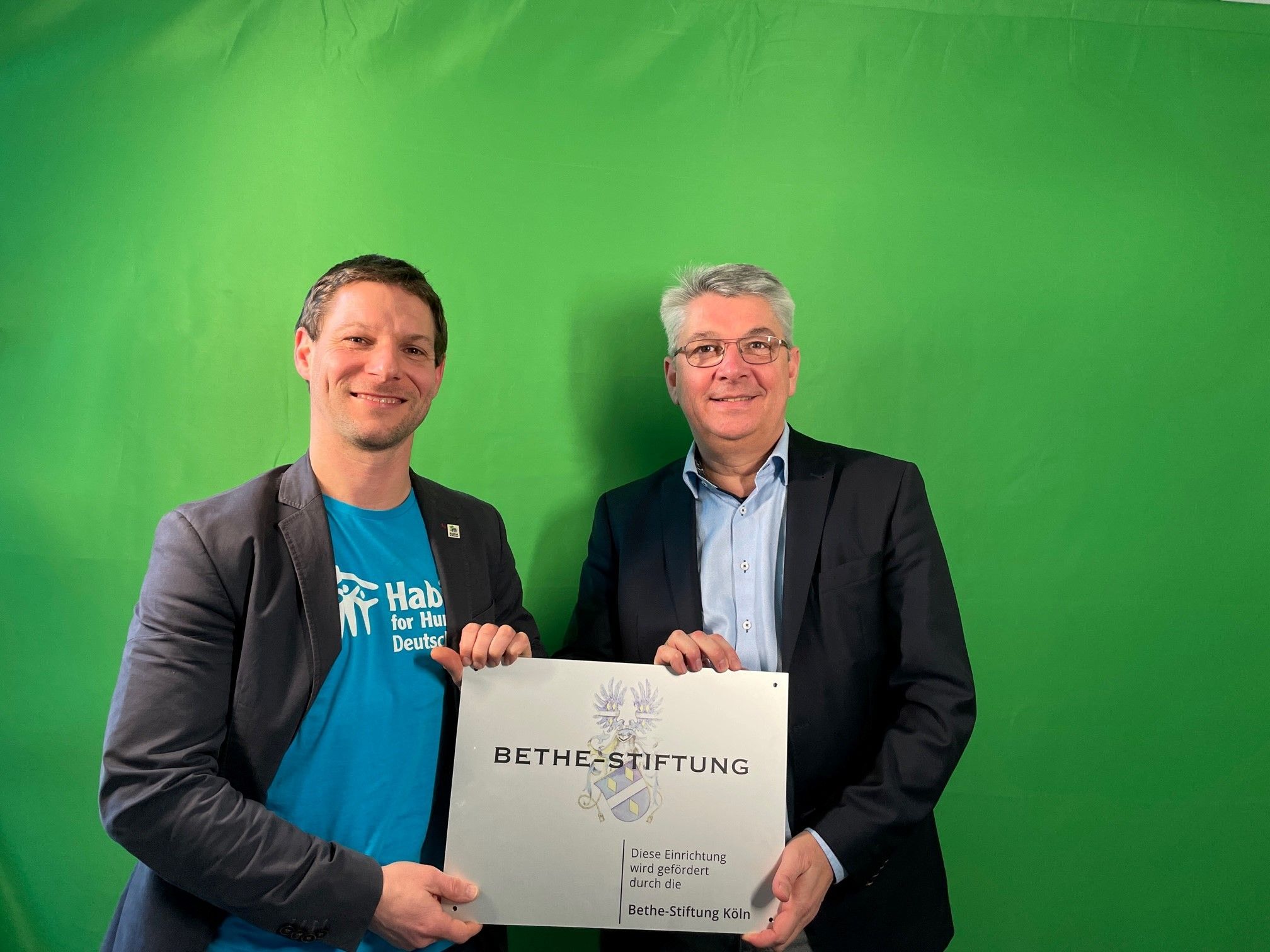 Der Vorstand der Organisation Habitat for Humanity, Gereon Fischer (links), und Lutz Urbach von der Bethe-Stiftung halten ein Schild der Bethe-Stiftung in Händen, das von der Stiftung geförderte Projekte kennzeichnet.