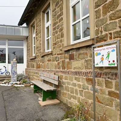 Ein altes Gebäude aus groben, unverputzten Steinquadern mit einem modernen Anbau. Davor eine Holzbank und ein buntes Kindergartenschild.