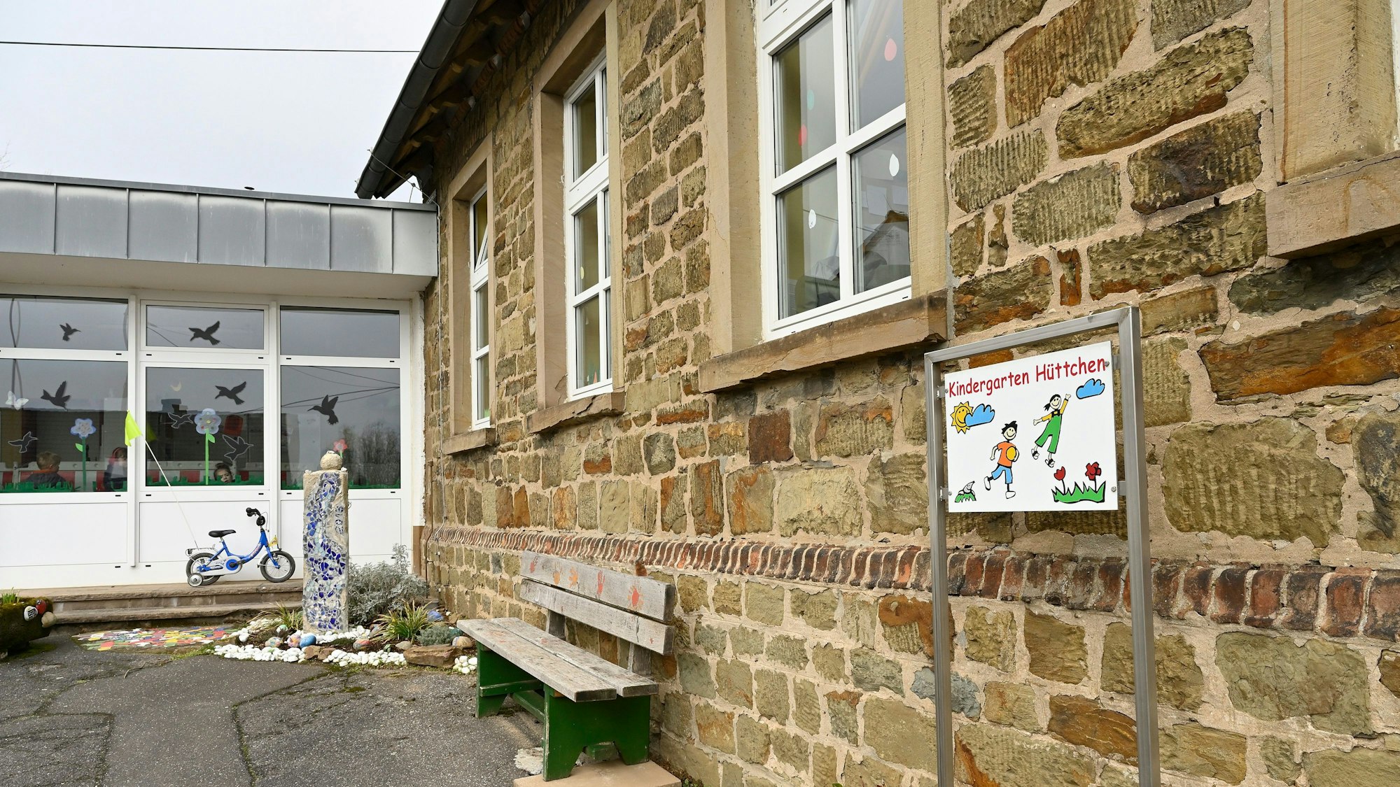 Ein altes Gebäude aus groben, unverputzten Steinquadern mit einem modernen Anbau. Davor eine Holzbank und ein buntes Kindergartenschild.