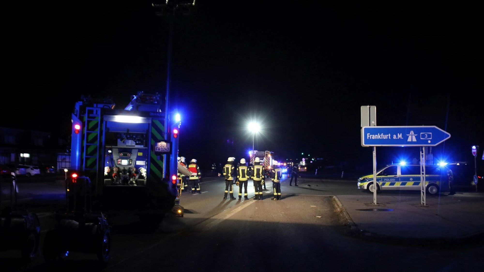 Feuerwehrleute stehen auf nächtlicher Straße auf einer ausgeleuchteten Straße neben dem Schild, das die Zufahrt zur Autobahn Richtung Frankfurt markiert.
