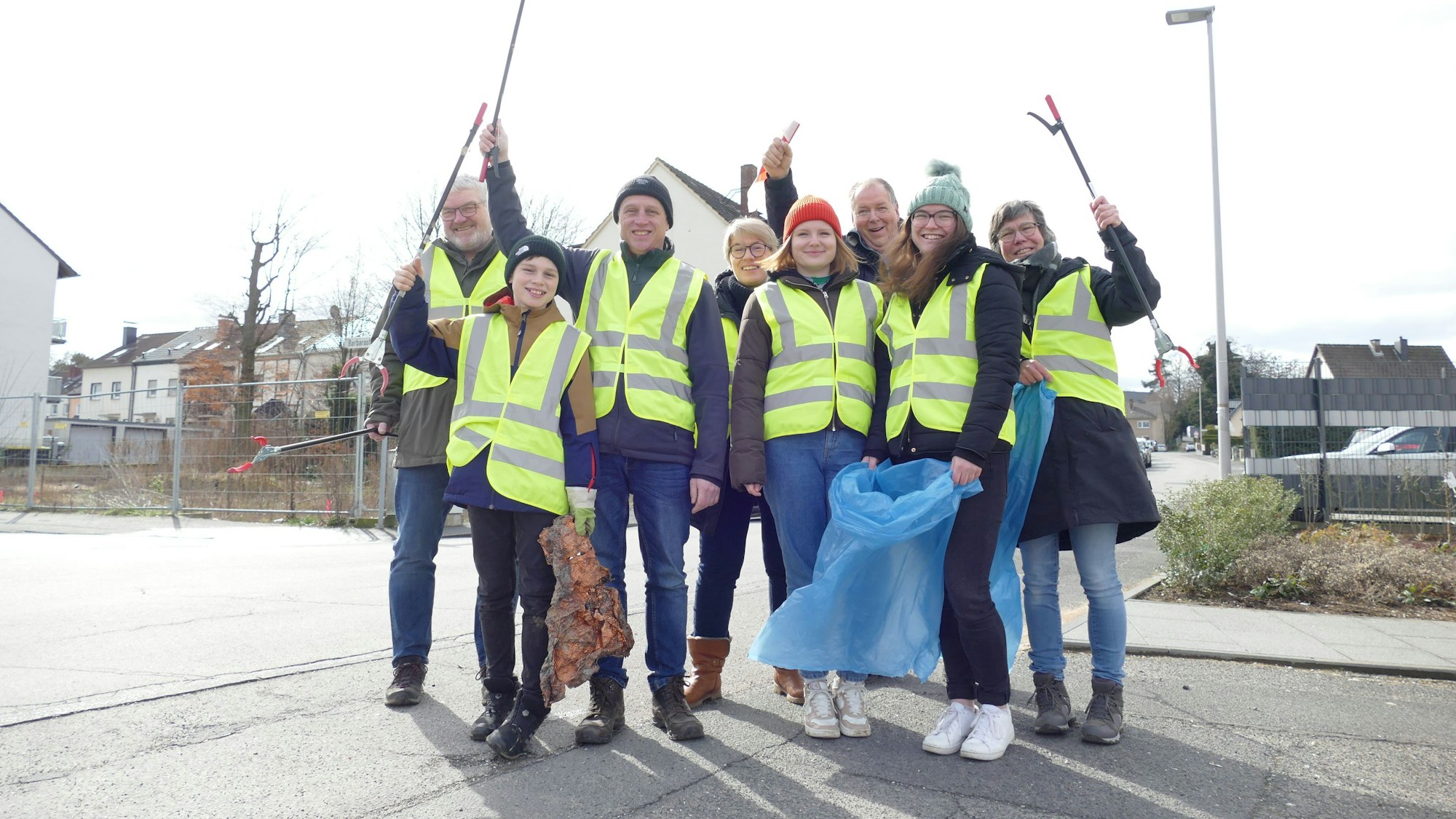Eine Gruppe Frauen, Männer und Kinder in gelben Warnwesten und mit Mülltüten schwenken ihre Greifzangen.