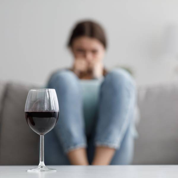 Im Hintergrund sitzt eine Frau auf einem Sofa, davor stehen eine leere Flasche Wein und ein volles Wein-Glas auf dem Tisch