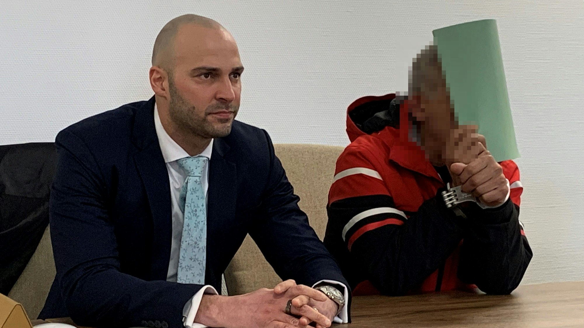 Man sieht den Angeklagten, der sich hinter einem Ordner verbirgt, neben ihm sein Verteidiger.