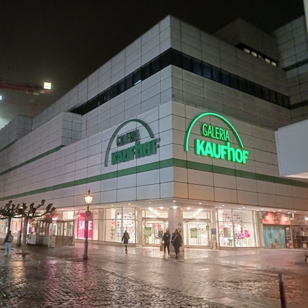 Die Front der Galeria Kaufhof an der Siegburger Kaiserstraße am Abend. Ein Schriftzug leuchtet, ein anderer ist dunkel.