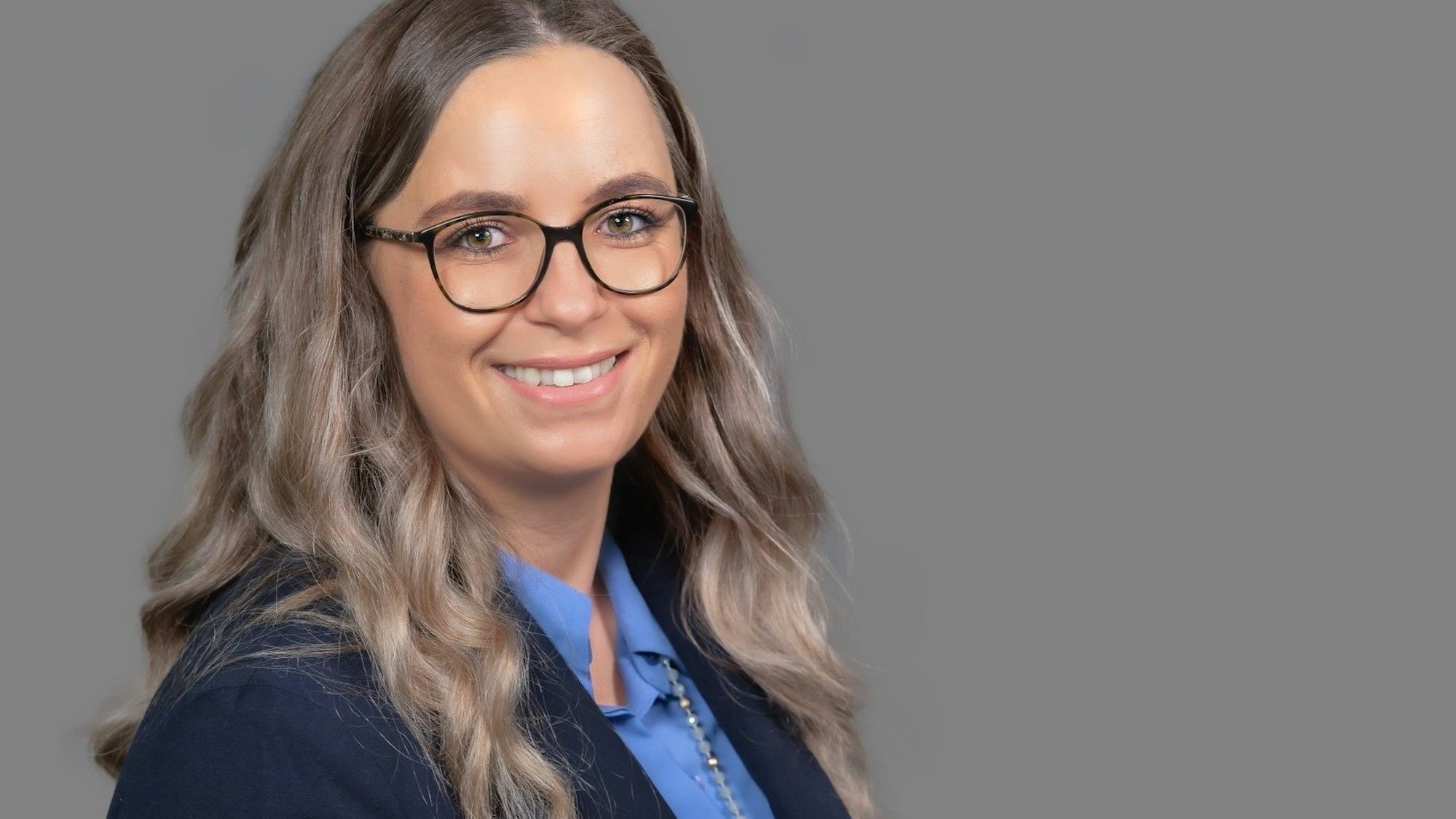 Frau Fox lächelt in die Kamera. Sie trägt eine Brille und ein blaues Hemd, mit dunkelblauem Blazer darüber.