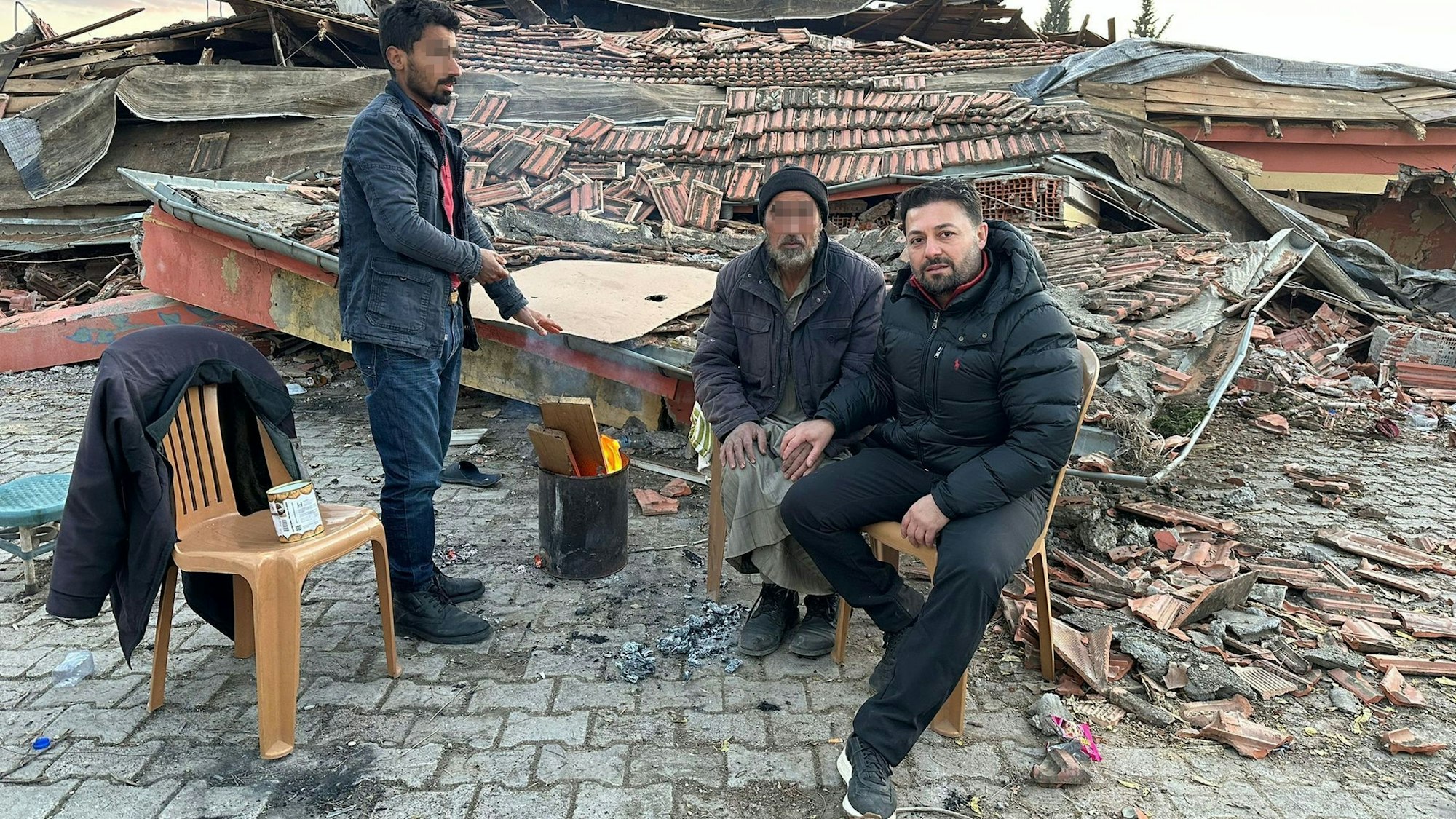 Der Euskirchener Ali Demirbas und zwei weitere Männer sitzen auf Plastikstühlen, um sie herum die Trümmer zerstörter Häuser.