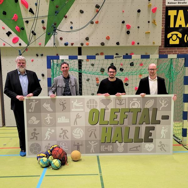 Die vier Männer stehen in einer Turnhalle und halten ein Plakat mit dem Namen in den Händen.