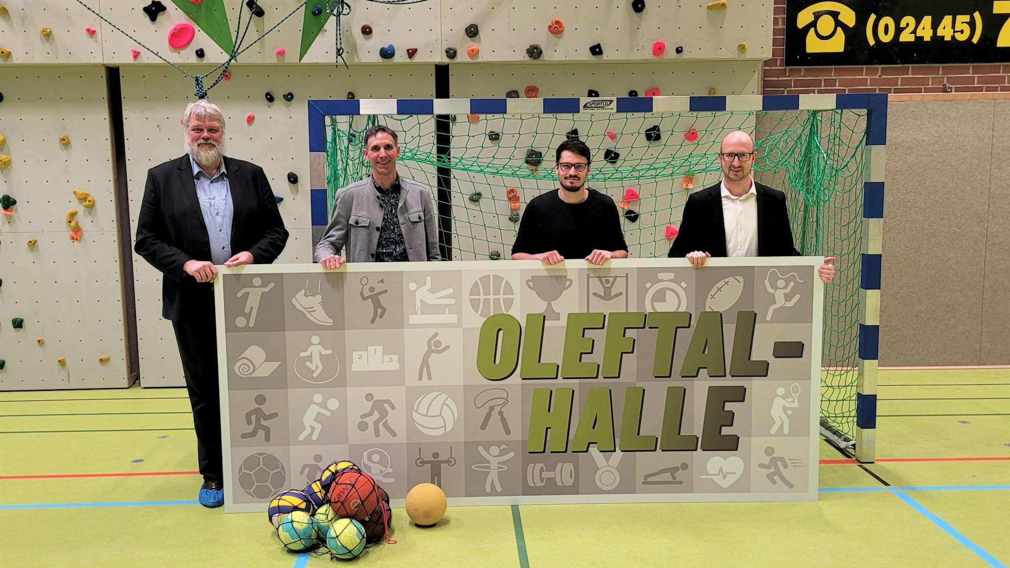 Die vier Männer stehen in einer Turnhalle und halten ein Plakat mit dem Namen in den Händen.