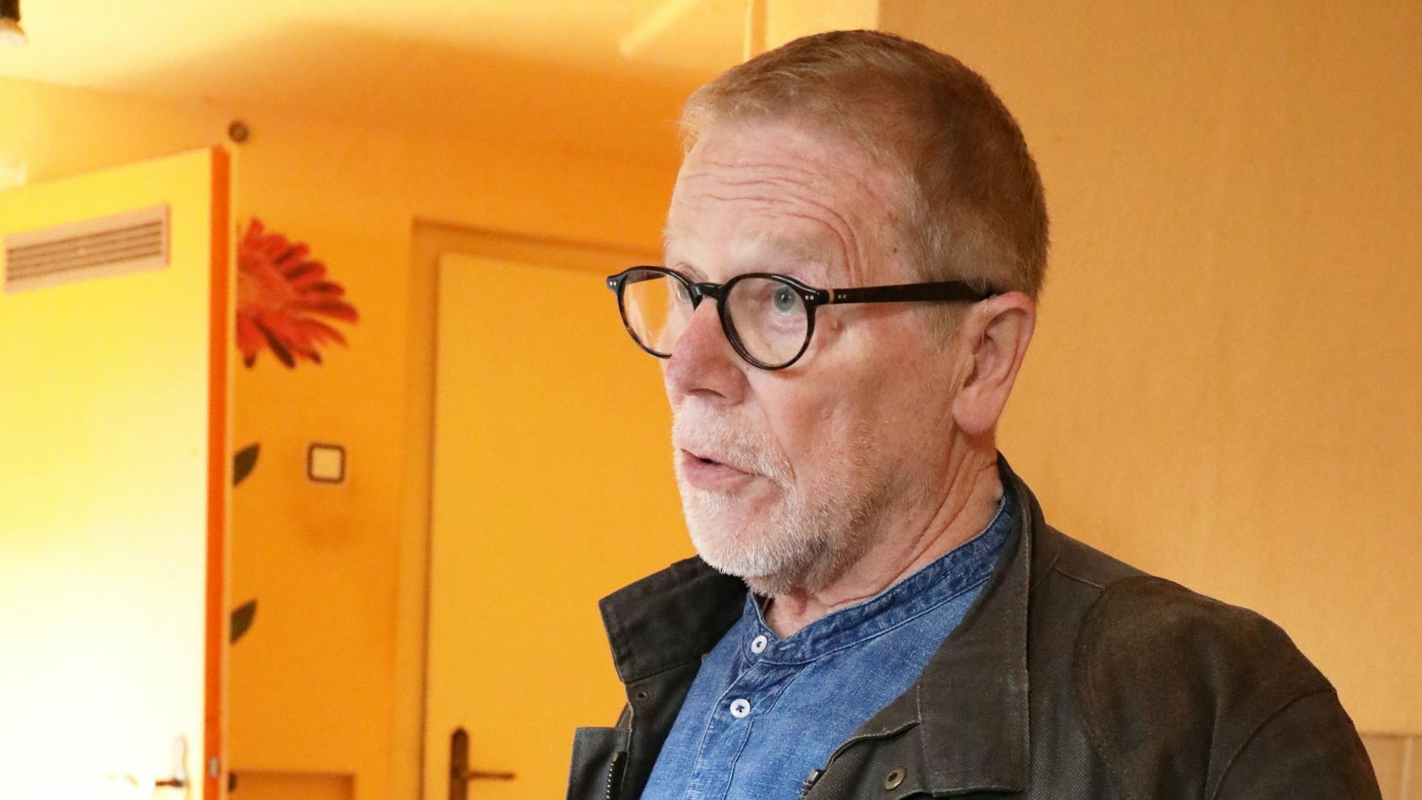 Ein Mann mit schwarzer Brille und blauem Hemd steht einem der Räume mit orange-gelben Wänden, die für eine Erweiterung im Gespräch sind.