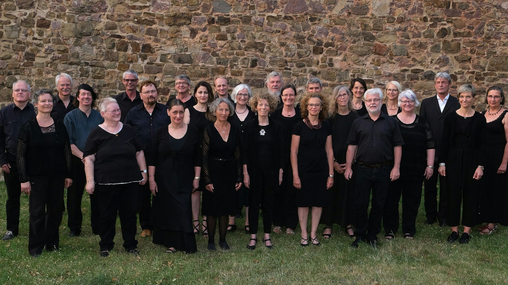 Etwa dreißig in schwarz gekleidete Personen stehen in drei Reihen aufgebaut vor einer Mauer.