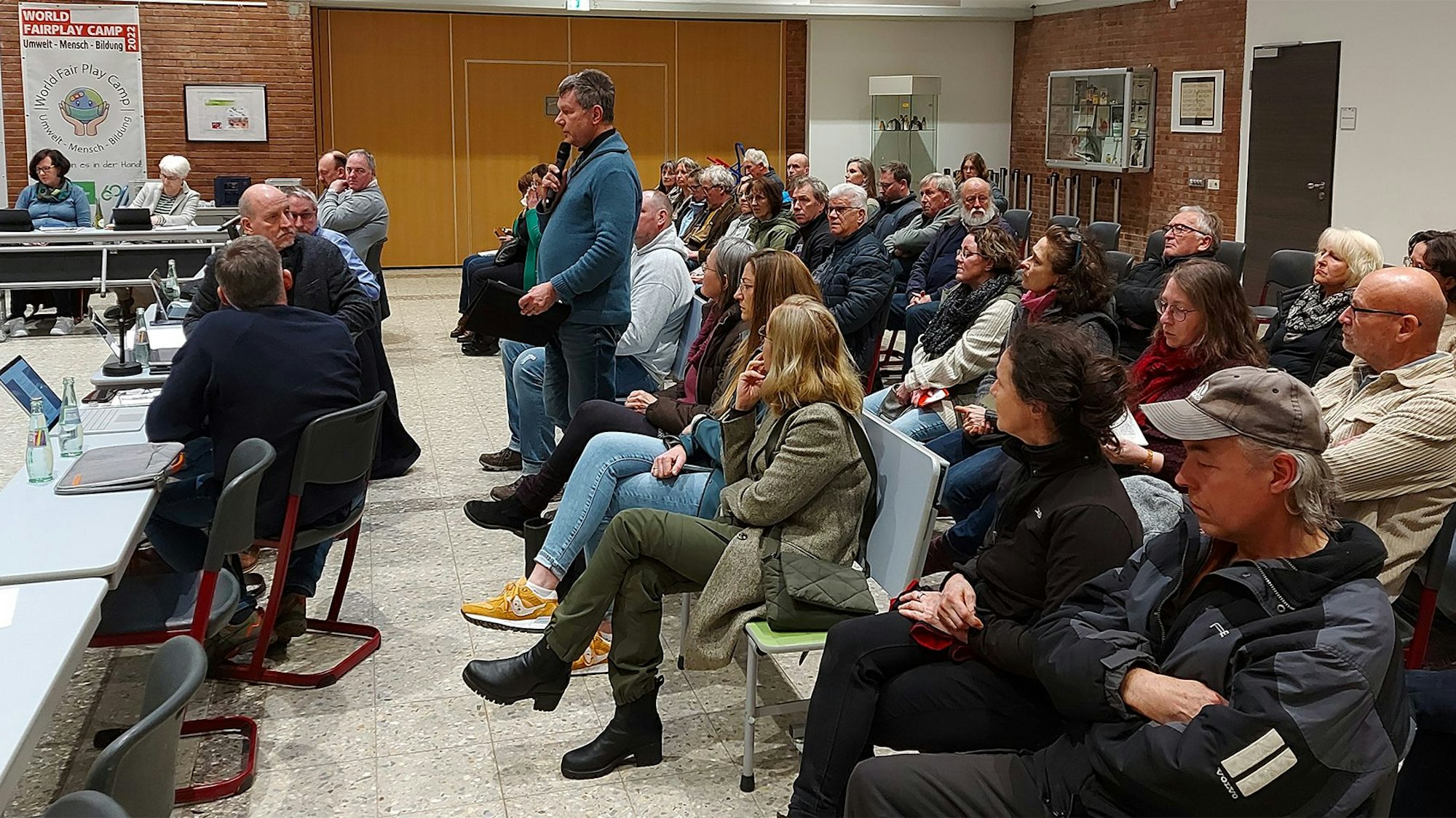 Ein Mann steht inmitten von vielen sitzenden Menschen in einer Aula.