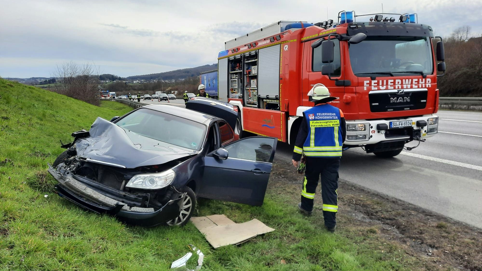Ein blauer Pkw mit stark verbeulter Front liegt auf einem Grünstreifen neben der Autobahn. Dahinter steht ein Einsatzfahrzeug der Feuerwehr.