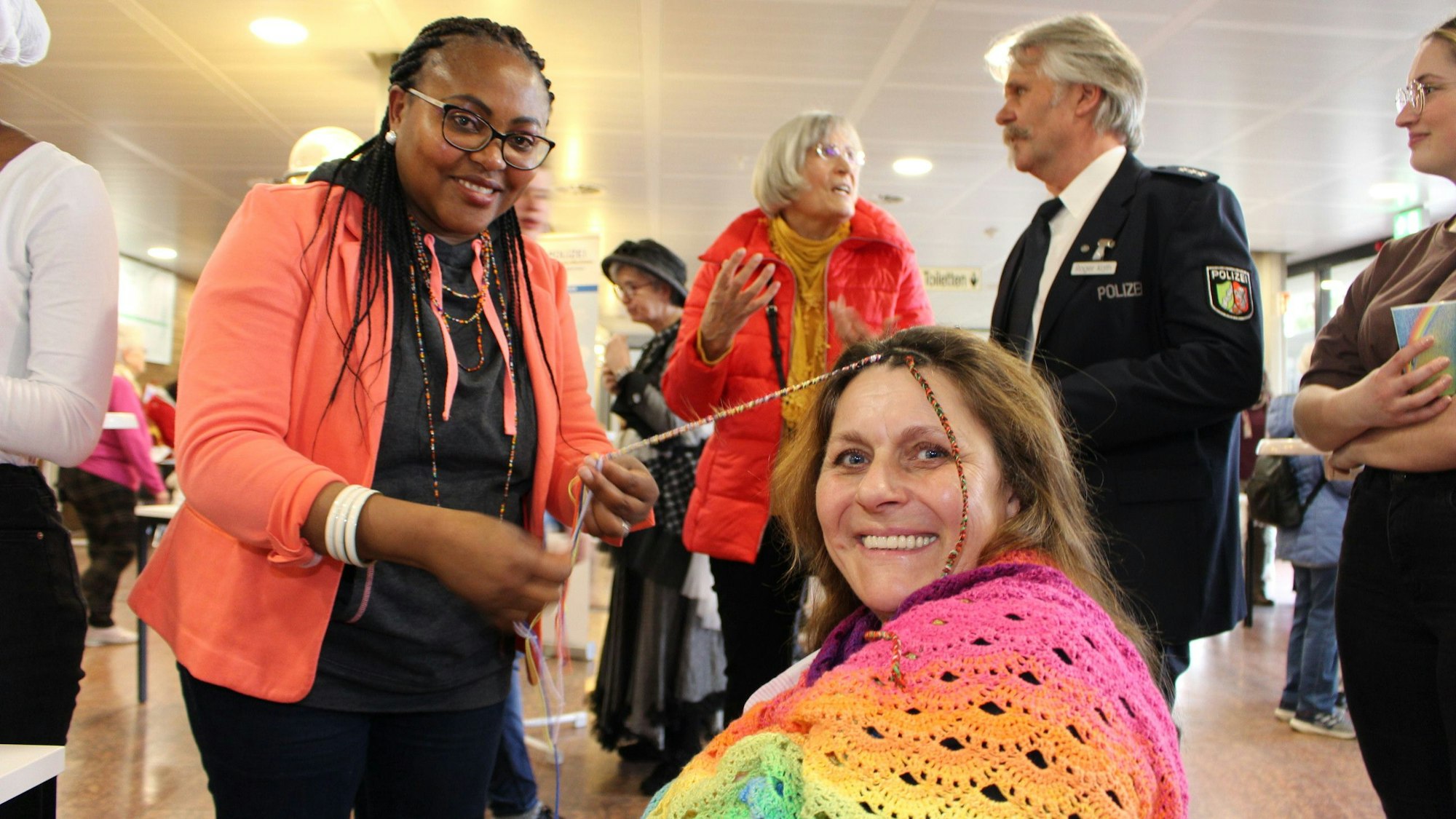 Epiphana Uwimana dreht einer in Regenbogenfarben gekleideten Besucherin bunte Fäden in die Haarsträhnen.