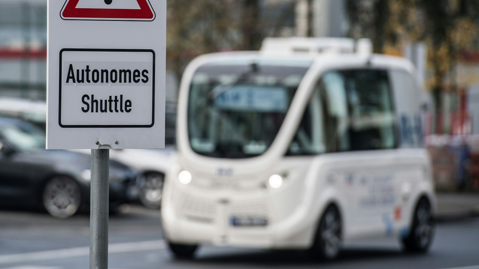 Ein Schild "Autonomes Shuttle" weist auf dem Frankfurter Flughafen auf den Testbetrieb mit selbstfahrenden Shuttles hin, das im Hintergrund fährt.