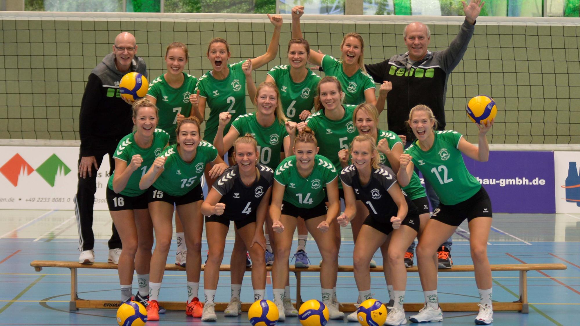 Volleyballerinnen des TVA Hürth haben sich zum Mannschaftsfoto aufgestellt und jubeln.