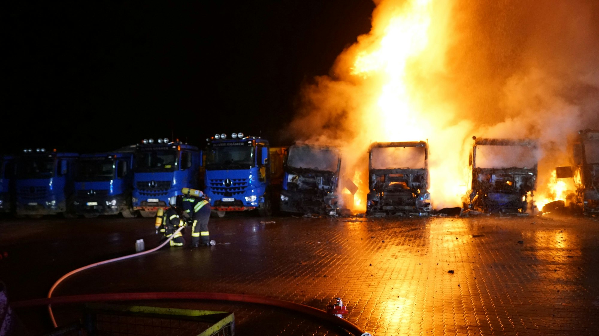 Dicht an dicht sind die Lastüge der Spedition geparkt. Man erkennt, dass drei der Laster lichterloh in Flammen anstehen. Auch an einem vierten Lkw ist Feuer zu sehen.