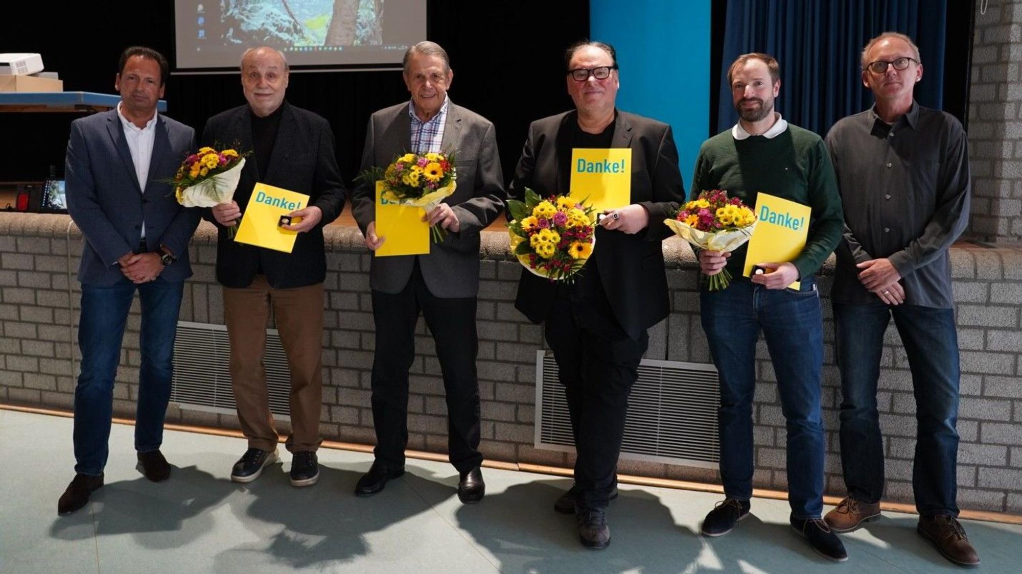 Mehrere Männer stehen mit Blumensträußen und Urkunden, auf denen „Danke!“ zu lesen ist, nebeneinander, zwei weitere gratulieren ihnen.
