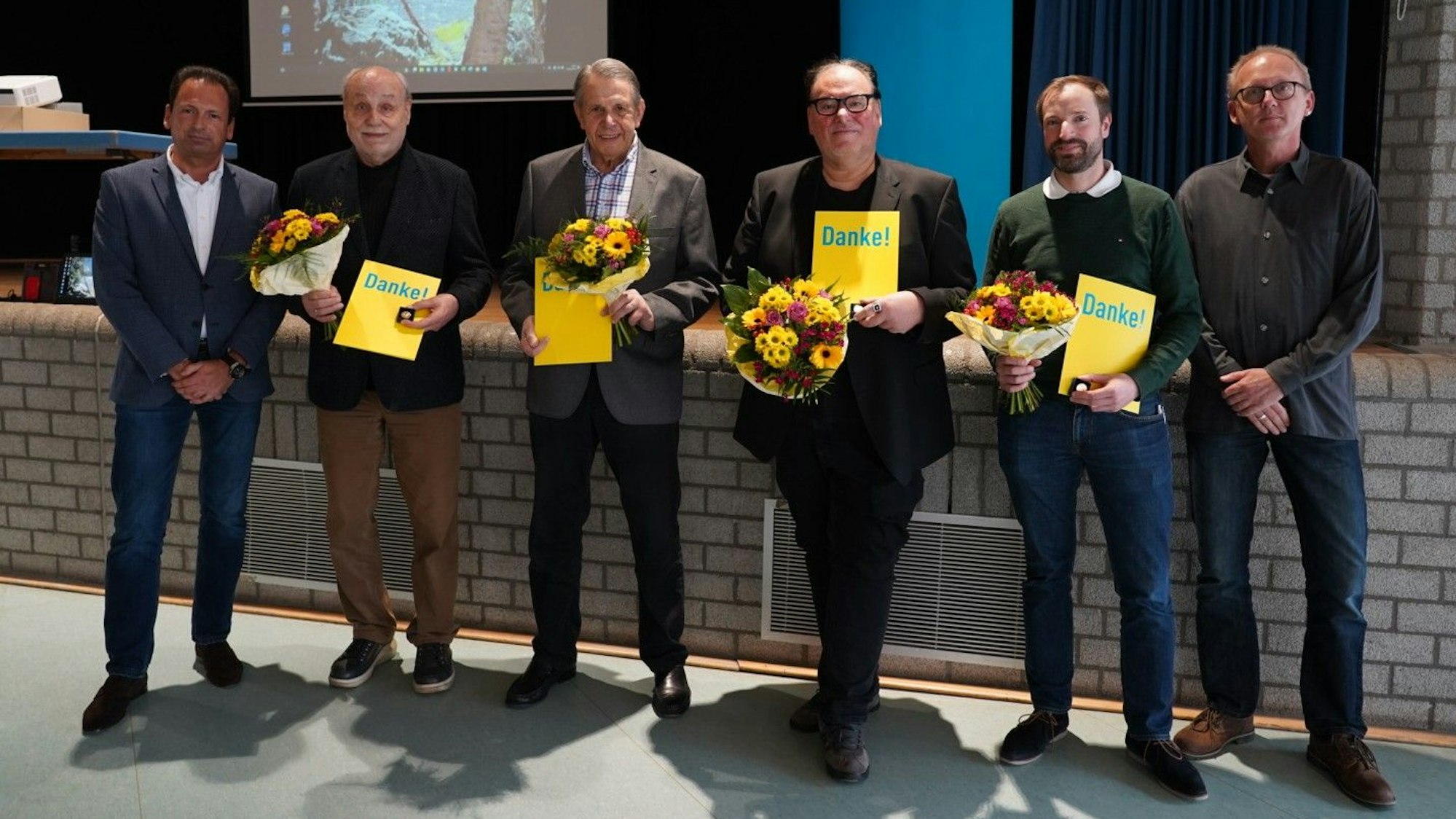 Mehrere Männer stehen mit Blumensträußen und Urkunden, auf denen „Danke!“ zu lesen ist, nebeneinander, zwei weitere gratulieren ihnen.
