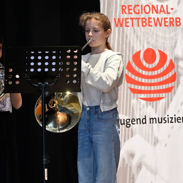 Die beiden stehen auf einer Bühne mit ihren Instrumenten, links im Bild ein Banner.