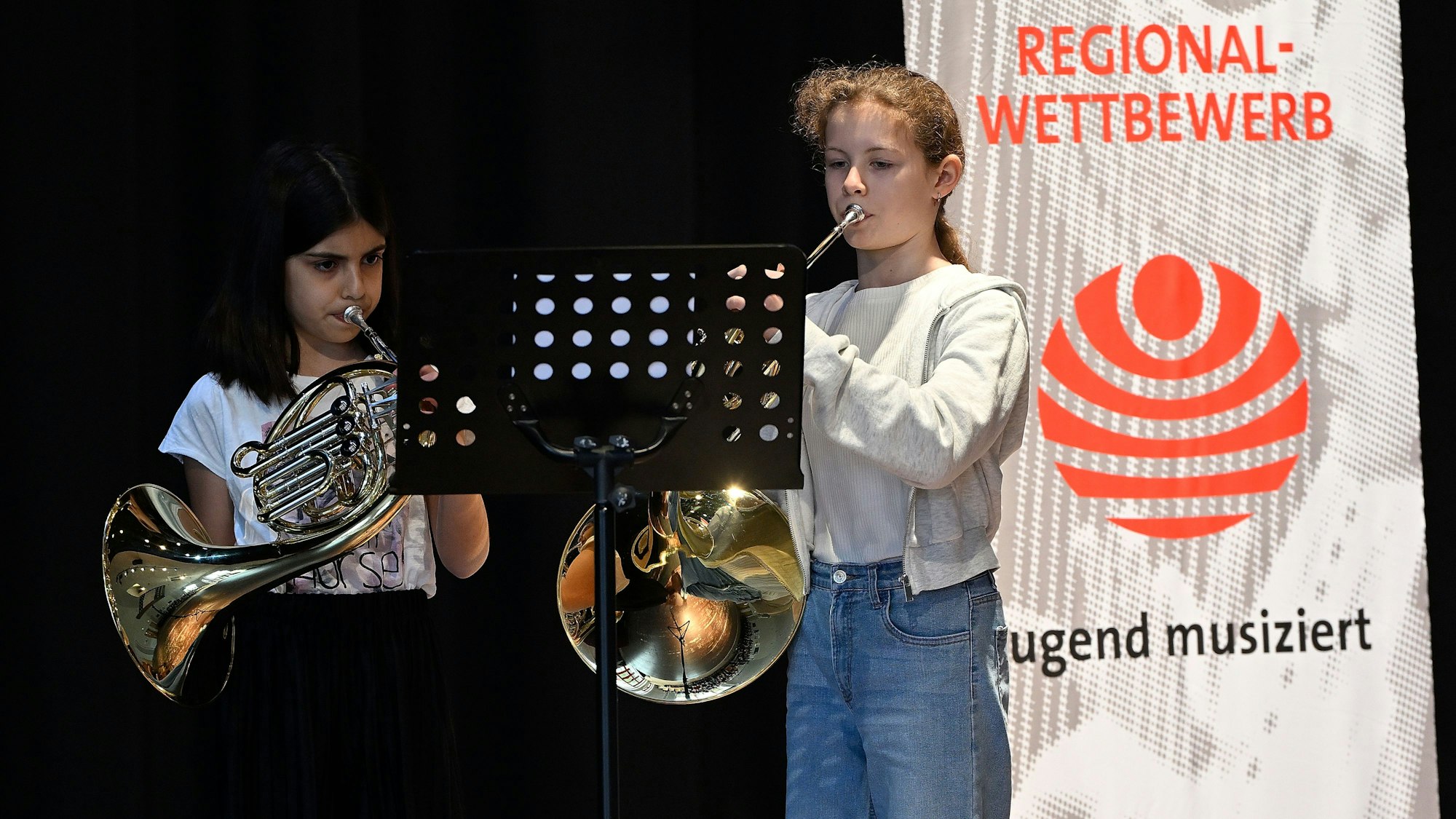 Die beiden stehen auf einer Bühne mit ihren Instrumenten, links im Bild ein Banner.