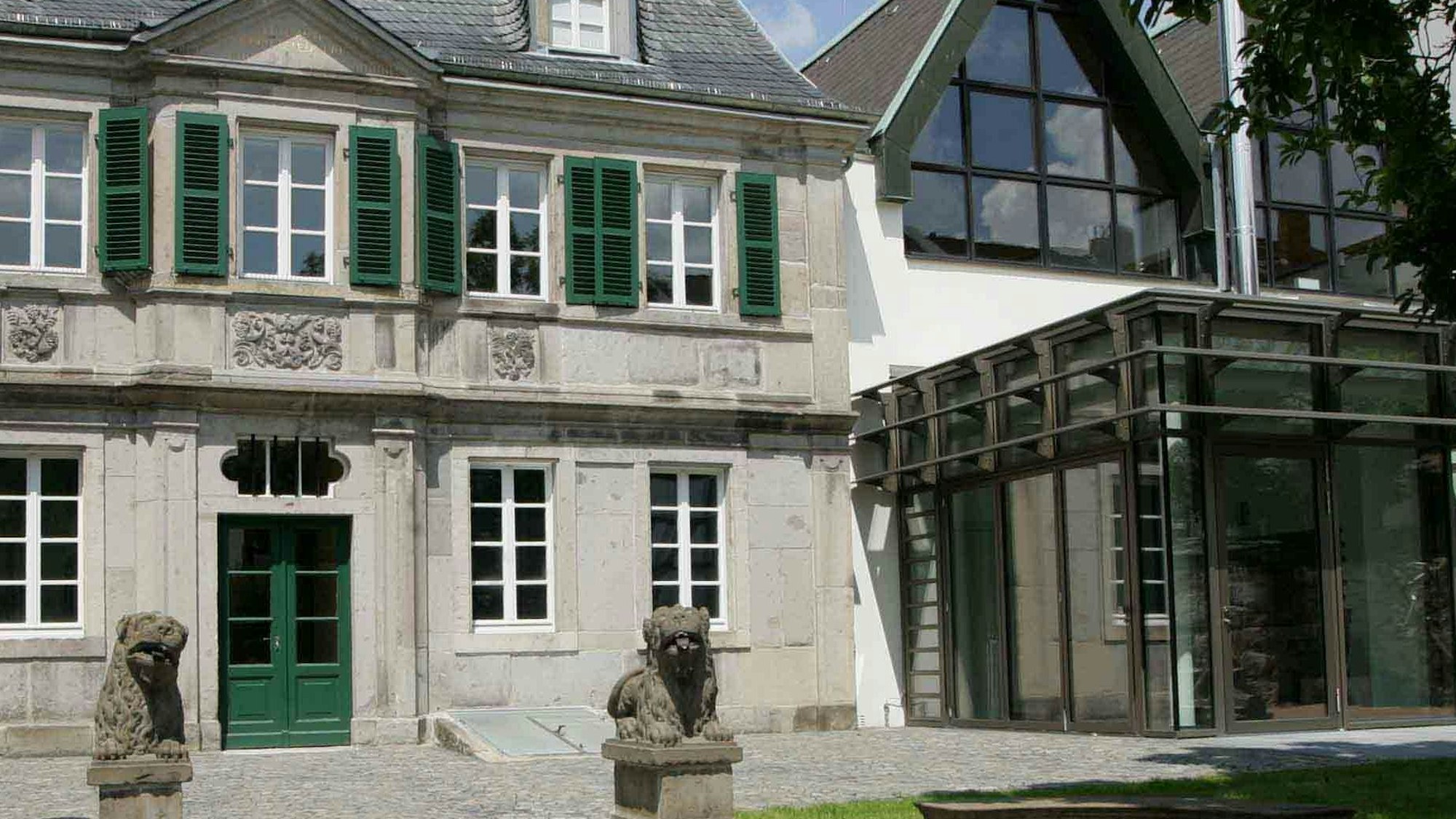 Zwei Steinlöwen sitzten auf Säulen vor einem Barockgebäude, dessen Tür und Fensterläden grün angestrichen sind. Rechs daneben befindet sich ein moderner Glasbau, um den das Museum erweitert worden ist.