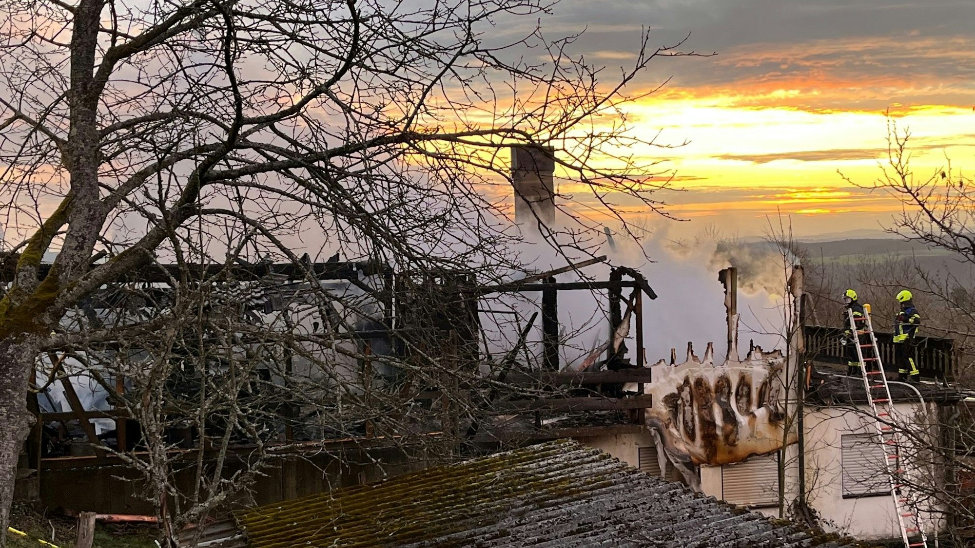 Rauch kommt aus den Überresten des Hauses. Am Bildrand sind Feuerwehrleute zu sehen.
