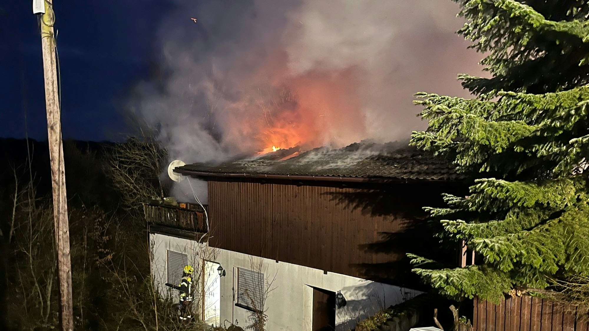 Auf dem Dach des Hauses sind Flammen zu sehen, vor der Tür steht eine Einsatzkraft der Feuerwehr.