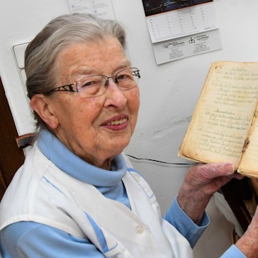 Oma Agnes sitzt an ihrem Küchentisch und zeigt ihr Rezeptbuch. Auf der Tischplatte liegen ausgerollter Teig und ein Nudelholz.
