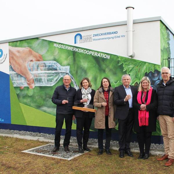 SIeben Personen stehen vor einem Gebäude und halten Plastikbecher mit Wasser in der Hand. Eine bemalte Hauswand im Hintergrund weist auf die Wasserwerkskooperation hin.