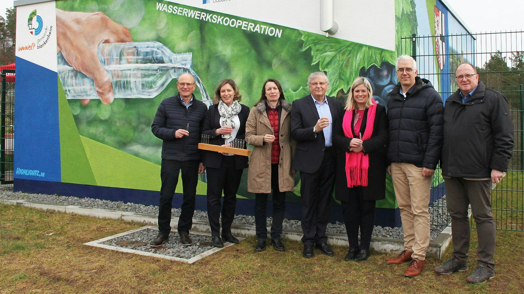 SIeben Personen stehen vor einem Gebäude und halten Plastikbecher mit Wasser in der Hand. Eine bemalte Hauswand im Hintergrund weist auf die Wasserwerkskooperation hin.