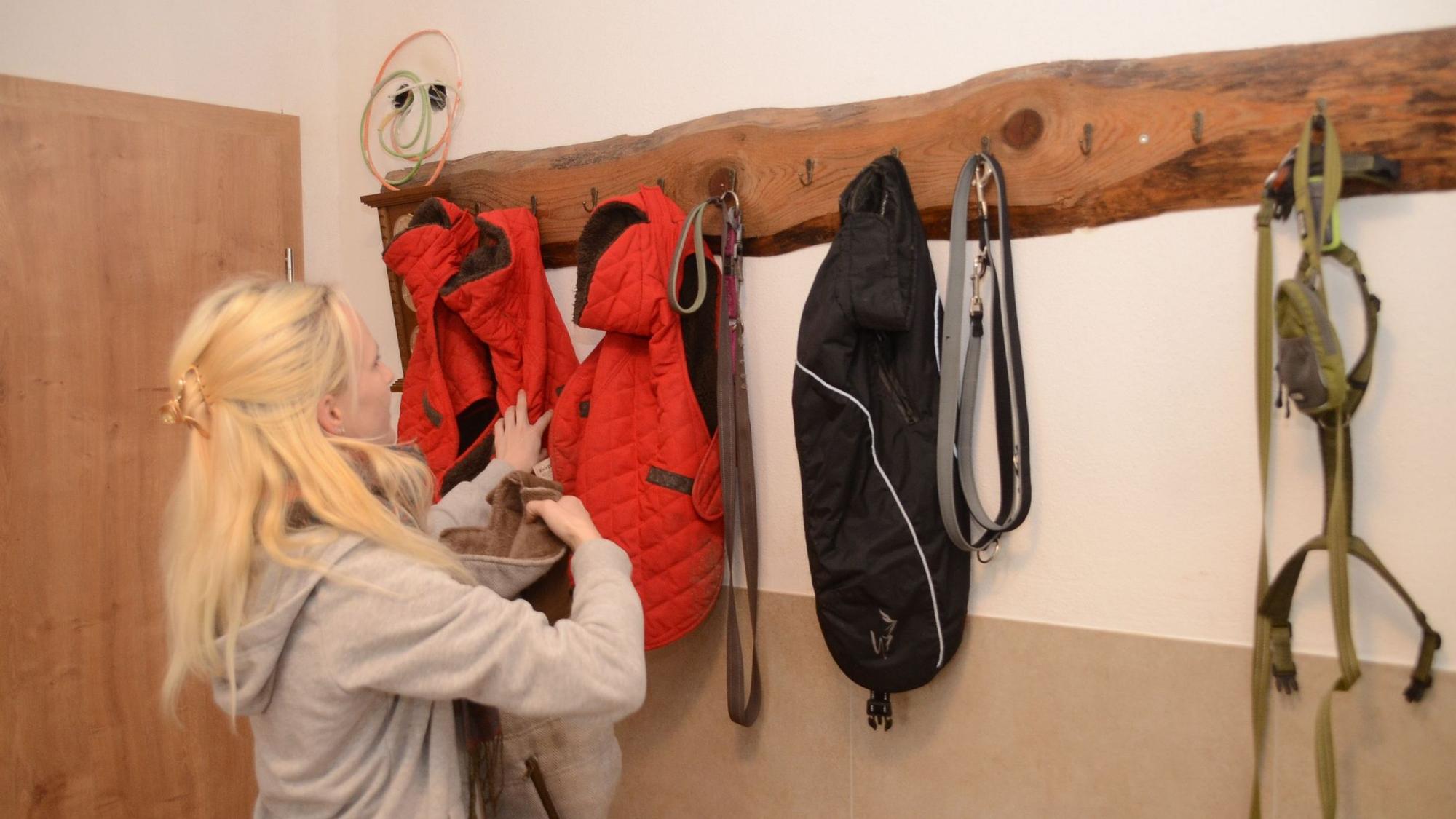 EIne blonde Frau hängt Hundemäntel und Leinen an einer Garderobe auf.